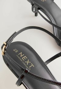 Sorte stroppandaler med et elegant design, blank finish, guldfarvet hardware detaljer og mærkenavnet "NEXT" i guld på indersålen.