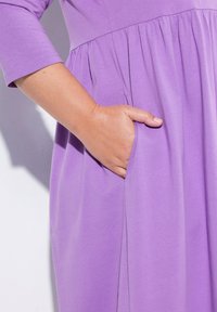 Violetti mekko, jossa on kolmasosa hihat, kerätty vyötärö ja sivutaskut. Pehmeä kankaan rakenne ja sileä pinta.