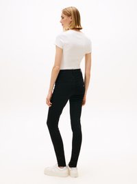 Kvinna med axellångt blont hår iförd en vit t-shirt med kort ärm, svarta slim-fit jeans och vita platåskor, stående i profil.