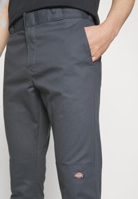 Szare spodnie robocze wykonane z wytrzymałej tkaniny, posiadające boczną kieszeń, przeszywane szwy oraz naszywkę z logo na lewym udzie.