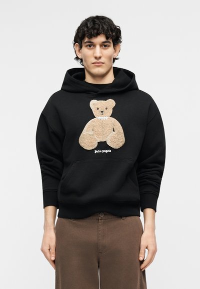 Jonge man draagt een zwarte hoodie met een beige teddybeerontwerp en de tekst "Palm Angels", gecombineerd met een bruine broek, staand tegen een witte achtergrond.