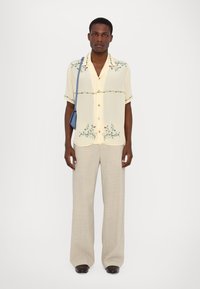 Camisa de manga corta de color amarillo claro con bordado floral, combinada con pantalones de pierna ancha beige y zapatos negros, sosteniendo un bolso de piel azul.