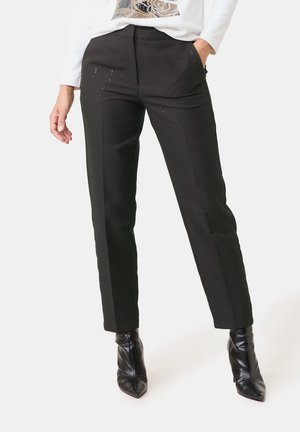 Donna che indossa pantaloni neri a gamba dritta con trama e stivaletti neri con punta, mano in tasca, top bianco parzialmente visibile.