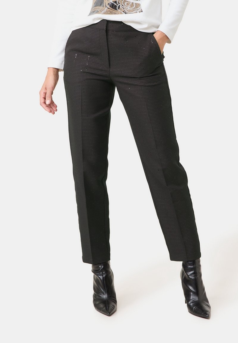 Femme portant un pantalon noir texturé à jambes droites et des bottines noires pointues, main dans la poche, haut blanc partiellement visible.