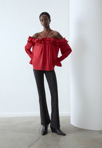 Blouse rouge décolletée sur les épaules avec volants, manches longues et poignets élastiques, associée à un jean noir évasé et des escarpins noirs pointus.