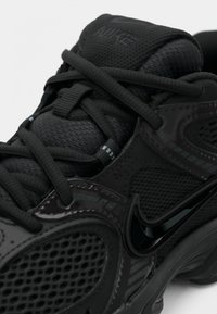 Μαύρο αθλητικό παπούτσι με δίχτυ και συνθετικά υλικά, που διαθέτει αντιθετικές υφές, έντονα κορδόνια και λαμπερό μαύρο λογότυπο Swoosh.