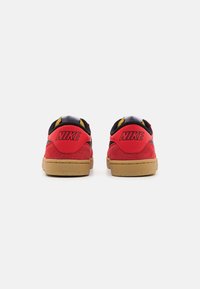 Baskets en daim rouge avec des accents noirs et des détails jaunes. Elles présentent une semelle texturée, des coutures visibles et un logo Nike bien en vue sur le talon.