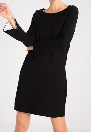 Femme portant une robe noire longueur genoux avec de longues manches cloche laissant apparaître une doublure intérieure blanche sur un poignet.