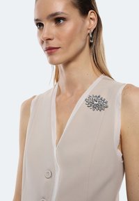 Top senza maniche beige con scollatura a V profonda, caratterizzato da una spilla decorativa argento sul lato sinistro. Il tessuto appare liscio e leggero.