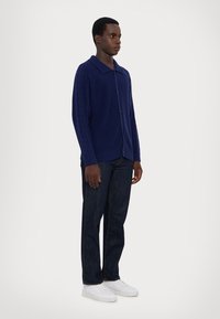 Navy knitted cardigan with a ribbed collar, front buttons, and cable knit texture on sleeves, paired with dark jeans and white sneakers.