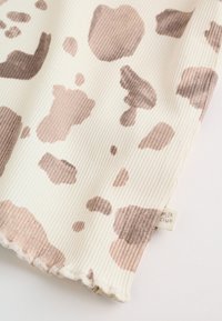 Tissu côtelé crème avec des taches irrégulières marron clair imprimées façon vache et une petite étiquette indiquant « milk club » près du bord festonné.
