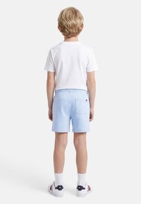 Παιδί με ξανθά μαλλιά, που φοράει λευκό μπλουζάκι, ανοιχτό μπλε σορτς, λευκές κάλτσες και λευκά αθλητικά παπούτσια, στέκεται με την πλάτη προς τα πίσω σε λευκό φόντο.