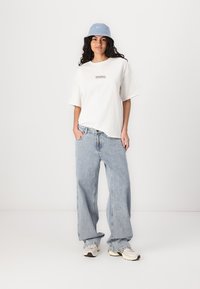 Camiseta blanca de manga corta con un pequeño gráfico, sombrero de cubo azul claro y jeans sueltos azul claro, combinados con zapatillas beige.