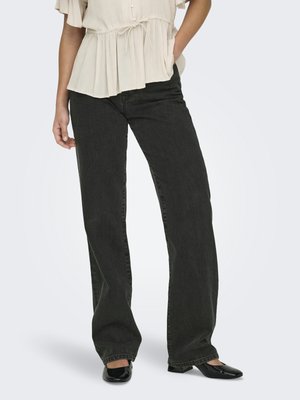 Person iført svarte vide jeans med høy midje, beige kortermet bluse og svarte sko med firkantet tå, stående med den ene hånden i lommen.