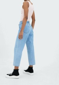 Henkilö, joka on pukeutunut vaaleansinisiin korkeavyötäröisiin farkkuihin, vaaleanpunaiseen topiin ja mustiin korkeus- sekä valkoisenpohjaisiin lenkkareihin, seisoo valkoisella lattialla.