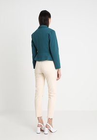 Tyrkysová peplum bunda s dlouhými rukávy, kombinovaná s krémově bílými slim kalhotami. Boty jsou bílé se širokým podpatkem a otevřenou patou.