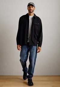 Mand går indendørs iført sort kasket, sort jakke, grå skjorte, blå jeans og sorte sko mod en ensfarvet baggrund.