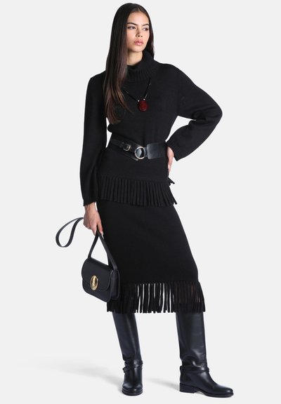 Femme portant un ensemble pull à franges et jupe noirs, large ceinture noire, bottes noires montant jusqu'au genou, tenant un sac à main noir avec une boucle dorée.