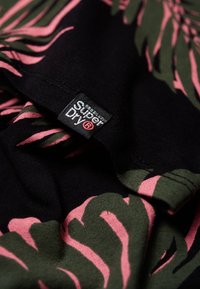 Tissu noir avec des motifs de feuilles verts et roses et une petite étiquette noire de la marque Superdry cousue dans la couture.