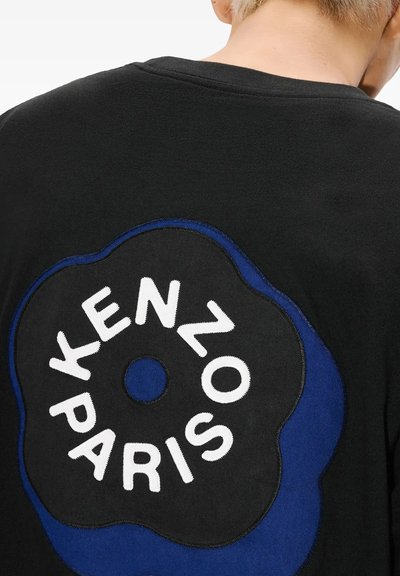 Person iført en sort skjorte med et stort rundt sort og blåt mærke på ryggen med hvid tekst, der lyder "KENZO PARIS."