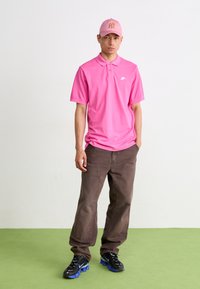 Rosa Polohemd mit kurzen Ärmeln und Kragen, dunkelbraune Hose und schwarze Sneakers mit blauen Akzenten. Eine passende rosa Mütze rundet den Look ab.