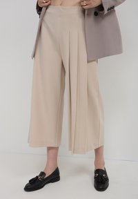 Beige weit geschnittene Hose mit vorderen Falten, kombiniert mit schwarzen Slippern mit Quasten und goldenen Akzenten. Der Stoff wirkt glatt und maßgeschneidert.