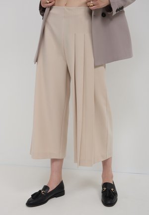 Beige weit geschnittene Hose mit vorderen Falten, kombiniert mit schwarzen Slippern mit Quasten und goldenen Akzenten. Der Stoff wirkt glatt und maßgeschneidert.