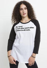 Biała koszulka baseballowa z czarnymi rękawami raglanowymi. Posiada czarny napis: "Bądź miły. Dbajcie o siebie. Słuchajcie alt rocka." Prosty design.