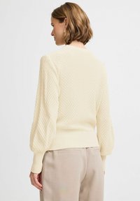 Pull en tricot crème avec un motif texturé, poignets et ourlet côtelés, manches ballons, porté sur un pantalon beige, vu de dos.