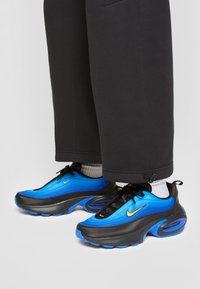 Person wearing black pants and white socks stands in blue and black Nike sneakers with yellow swoosh logos on light background.