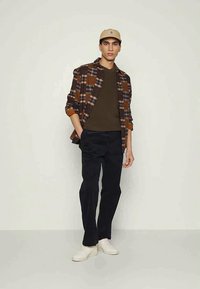 Camisa de flanela xadrez castanha sobre uma t-shirt verde escura, calças pretas e ténis brancos; usando um boné bege. Um conjunto simples e casual.