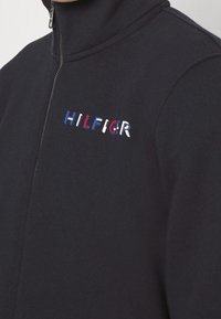 Giacca in pile blu navy con zip, con il logo "HILFIGER" ricamato in rosso, bianco e blu sul lato sinistro del petto. Texture liscia e colletto alto.