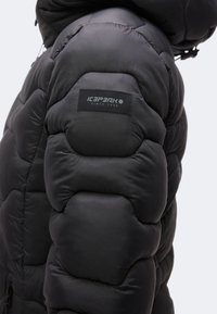 Manche de veste noire matelassée avec une étiquette de marque Icepeak cousue et des détails de couture visibles.