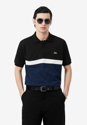 Joven con una camiseta polo negra, blanca y azul marino con un pequeño logo de cocodrilo, pantalones negros, cinturón negro, gafas de sol y una pulsera plateada.