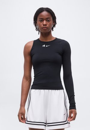 Žena oblečená v čiernom asymetrickom top s dlhým a krátkym rukávom a bielych atletických šortkách s čiernymi pruhmi.