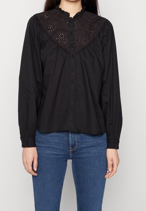 Kvinde iført en sort bluse med lange ærmer med knapper foran og øjetblonde-detaljer på bærestykket, kombineret med blå jeans.