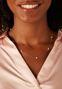 Zilveren ketting met verschillende kleine, ronde, lichtblauwe kralen die gelijkmatig over een delicate ketting zijn verdeeld, gedragen over een roze satijnen blouse.