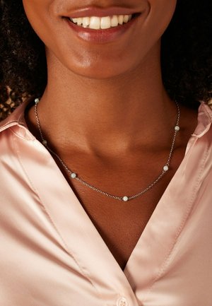 Zilveren ketting met verschillende kleine, ronde, lichtblauwe kralen die gelijkmatig over een delicate ketting zijn verdeeld, gedragen over een roze satijnen blouse.