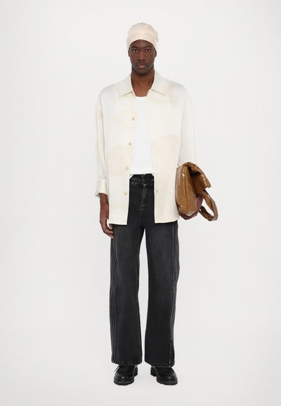 Camisa crema de gran tamaño con cierre de botones, combinada con jeans de pierna ancha negros y botas negras voluminosas. Sosteniendo una bolsa tote marrón.