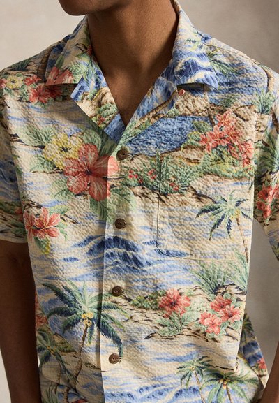 Muž nosí košeľu s krátkym rukávom na gombíky s tropickým kvetinovým a palmovým vzorom v modrých, zelených, červených a béžových tónoch.