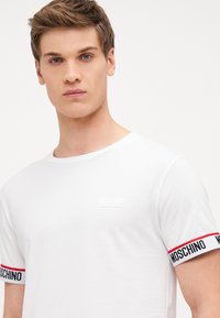 Biely bavlnený tričko s krátkymi rukávmi, s vyvýšeným logom Moschino na hrudi a kontrastnými pruhovanými akcentmi na manžetách rukávov.