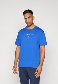 Blå kortærmet T-shirt lavet af bomuld, med teksten "PARIS 56" og et logo i hvid. Enkel design med rund halsudskæring.