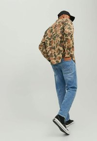 Camisa de camuflaje en tonos tierra, jeans de mezclilla azul claro y zapatos negros. La camisa tiene mangas largas y puños con botones. Lleva puesta una gorra sencilla.