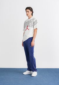 Fehér grafikai póló fekete és piros mintával, kék jogger nadrággal, fehér oldalsávokkal, és fehér sportcipővel párosítva.