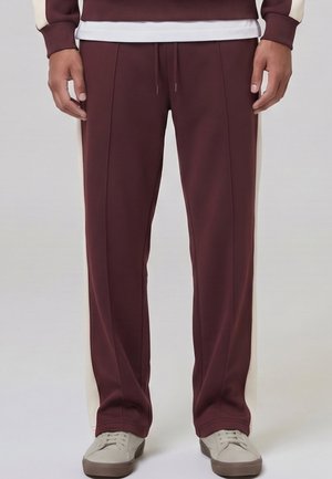 Homme portant un pantalon de survêtement bordeaux avec des bandes blanches sur les côtés et des baskets beiges, debout devant un fond uni.