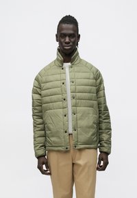 Muž v olivovo zelenej prešívanej bunde a béžových nohaviciach stojí pred jednoduchým bielym pozadím.