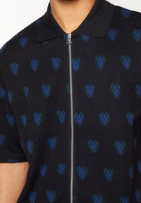 Polo shirt nero a maniche corte con zip frontale, decorato con un motivo di cuori verdi e blu. Tessuto morbido con un design del colletto classico.