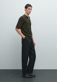 Polo shirt verde scuro a maniche corte, pantaloni neri larghi e sandali neri con design a tagli, su uno sfondo neutro.