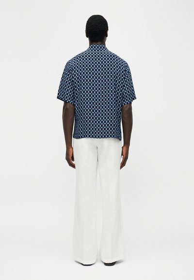 Moški stoji obrnjeno stran, oblečen v temnomodro srajco s kratkimi rokavi z svetlomodrim okroglim vzorcem in bele široke hlače.