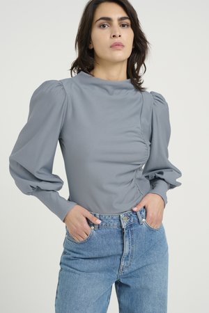Grå topp med puffede ermer i et glat stoff, med høy hals og samlet midje, kombinert med høyhalsede blå denimjeans.